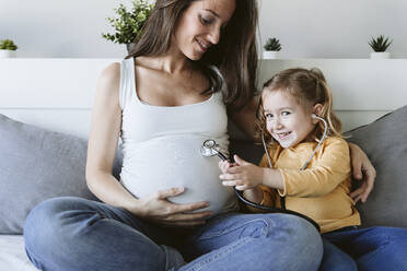 Lächelnde Tochter mit Stethoskop von schwangerer Mutter auf Bett zu Hause - EBBF05222
