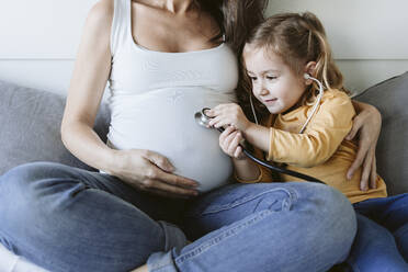 Girl listening to pregnant mother's belly through stethoscope at home - EBBF05223