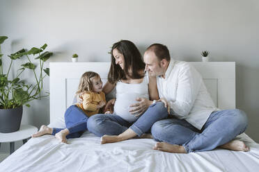 Glücklicher Mann und Frau betrachten ihre Tochter, die zu Hause auf dem Bett sitzt - EBBF05233