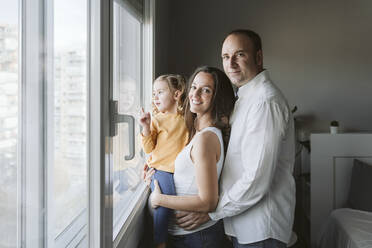 Smiling parents standing with daughter looking through window at home - EBBF05242