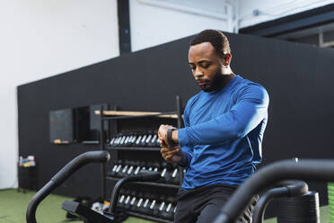 Athlete using fitness tracker sitting on airbike in gym - PNAF02878