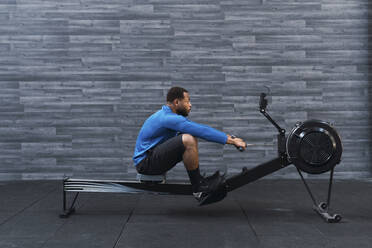 Concentrated athlete exercising on rowing machine in front of wall in gym - PNAF02879