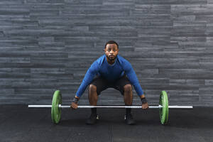 Konzentrierter junger Sportler, der eine Langhantel vor einer Wand im Fitnessstudio aufhebt - PNAF02895