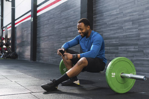 Lächelnder Sportler, der ein Smartphone benutzt und auf einer Hantel im Fitnessstudio sitzt - PNAF02896