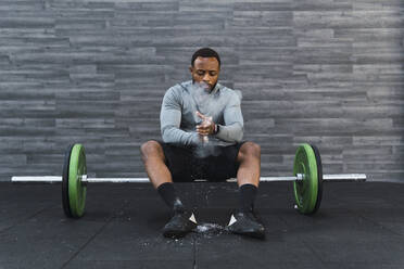 Sportler beim Auftragen von Kreidepulver, sitzend auf einer Langhantel im Fitnessstudio - PNAF02918