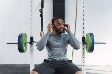 Nachdenklicher Sportler beim Einstellen der Kopfhörer auf einer Bank im Fitnessstudio - PNAF02928