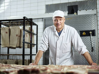 Smiling butcher wearing cap leaning on table in factory - CVF01865