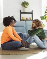 Seitenansicht von positiven multirassischen Freundinnen mit lockigem Haar, die sich entspannen, während sie auf einem Weidenteppich auf dem Boden im Wohnzimmer sitzen und sich gegenseitig anschauen - ADSF33437