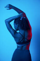 Seductive African American female model wearing black top with bare  shoulders holding raised arm in front and looking at camera during photo  session in dark studio stock photo