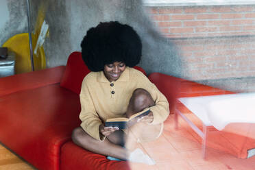 Front view of young African American female with Afro hairstyle framed between the reflections of a terrace while reading book while sitting legs crossed on comfortable red couch - ADSF33452