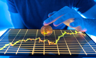 Crop anonyme männliche hält bitcoin Münze auf dem Display des Tablet mit finanziellen Chart auf dem Bildschirm in Neonlicht - ADSF33463