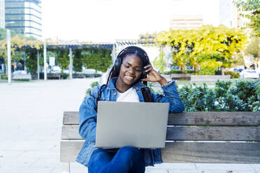 Junge Frau lächelt, während sie mit Laptop und Kopfhörern auf einer Bank in der Stadt sitzt - RFTF00155