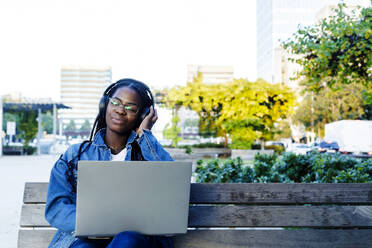 Junge Frau lächelt, während sie mit Laptop und Kopfhörern auf einer Bank in der Stadt sitzt - RFTF00156