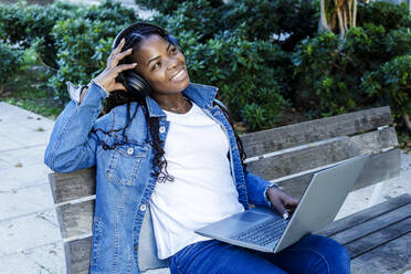 Young woman smiling while sitting on city bench with laptop and headphones - RFTF00157