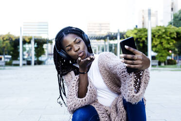 Junge Frau mit Kopfhörern hockt und bläst einen Kuss auf ihr Smartphone - RFTF00158