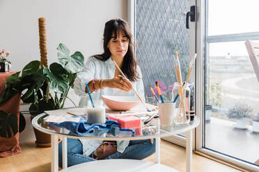 Concentrated talented female artist painting ceramic bowl while sitting at table with set of paintbrushes in light studio near door - ADSF33468