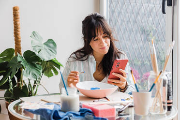 Konzentrierte Frau surft auf einem modernen Handy, während sie eine Schale am Tisch mit verschiedenen Pinseln in einer hellen, modernen Werkstatt mit Grünpflanzen bemalt - ADSF33469