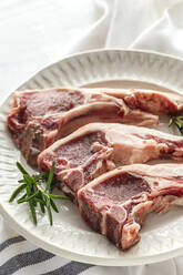 From above of uncooked lamb chops served on white ceramic plate with fresh green rosemary on table in light kitchen - ADSF33494