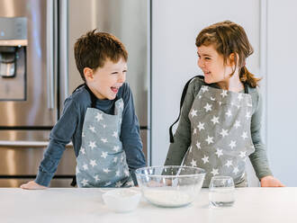 Happy little girl and boy in aprons standing on kitchen near table with glass bowl of flour and looking at each other - ADSF33497