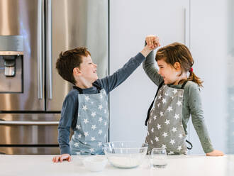 Happy little girl and boy in aprons standing on kitchen near table with glass bowl of flour and looking at each other holding hands - ADSF33498