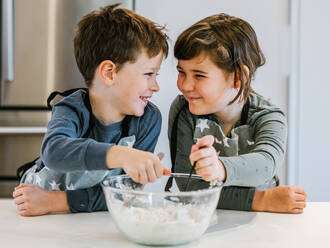 Happy little kids in aprons stirring flour in glass bowl while preparing dough and cooking together in kitchen at home and looking at each other - ADSF33500