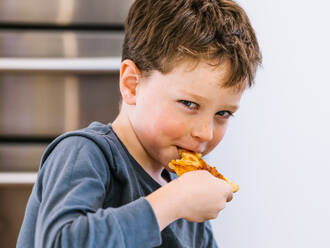 Happy little kid with brown hair eating piece of tasty homemade pizza while sitting in kitchen and looking at camera - ADSF33504