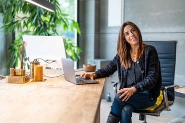 Fröhliche Frau in stilvollem schwarzem Outfit schaut in die Kamera, während sie an einem hölzernen Schreibtisch mit Computer in einem hellen modernen Arbeitsplatz sitzt - ADSF33506