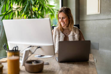 Fröhliche Arbeitnehmerin, die auf einem Laptop tippt, während sie auf einen Computermonitor in einem hellen, modernen Arbeitsbereich mit Grünpflanzen blickt - ADSF33508