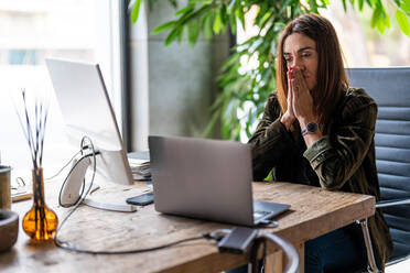 Worried female worker with folded hands looking at screen of laptop at wooden desktop during work in light modern office - ADSF33510