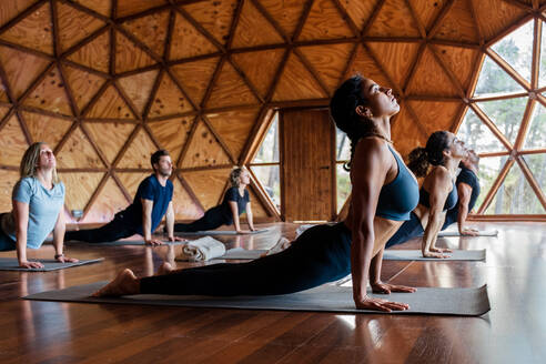 Gruppe aktiver Menschen in Sportkleidung, die die Bhujangasana-Haltung einnehmen, während sie während einer Yoga-Sitzung in einem hellen, geräumigen Studio auf Matten liegen - ADSF33515
