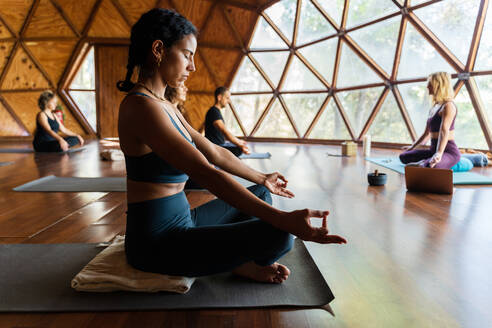 Menschen vor ihrem Lehrer in Sportkleidung, während sie in Padmasana-Pose sitzen, während sie Atemübungen während einer Yoga-Sitzung im Studio machen - ADSF33520