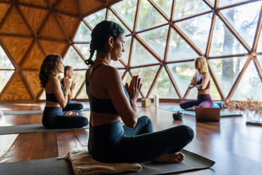 Menschen vor ihrem Lehrer in Sportkleidung, während sie in Lotus-Pose mit Namaste-Händen sitzen, während sie Atemübungen während einer Yoga-Sitzung im Studio machen - ADSF33521