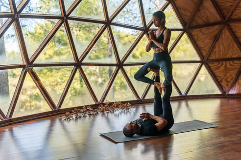 Ganzkörperaufnahme eines sportlichen Paares in Sportkleidung, das eine Yogapose einnimmt, während er sich mit ausgestreckten Beinen hinlegt und sie die Namaste-Pose einnimmt, während sie auf ihren Füßen in einem hellen Studio in der Nähe der Fenster sitzt - ADSF33522