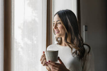 Smiling woman with coffee cup looking through window - EIF03197