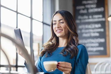 Lächelnde Frau mit Kaffeetasse bei einem Videogespräch über ein Mobiltelefon in einem Café - EIF03213