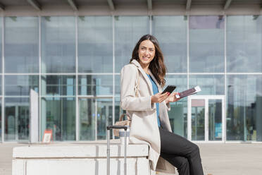 Smiling businesswoman with mobile phone and passport documents on bench - EIF03237