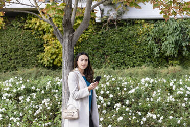Junge Geschäftsfrau mit Smartphone an einem Baum im Park - EIF03249