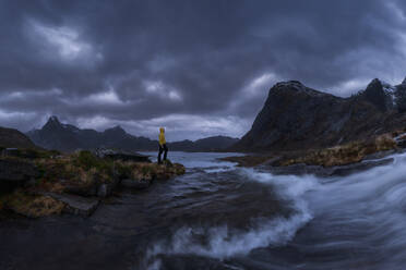 Tourist in yellow raincoat standing on grassy store near splashing water of Molneva Waterfall against gray overcast sky on stormy day on Lofoten Islands, Norway - ADSF33527
