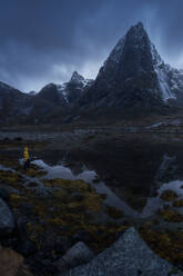 Tourist in yellow raincoat standing on grassy store near splashing water of Molneva Waterfall against gray overcast sky on stormy day on Lofoten Islands, Norway - ADSF33528