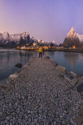 Stone quay located on calm sea water with tourist leading to Reine village and snowy mountain ridge against sundown sky on Lofoten Islands, Norway - ADSF33530