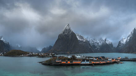 Das Dorf Reine liegt auf einer Insel, umgeben von ruhigem, blauem Wasser, schneebedeckten Bergen und bewölktem Himmel bei Sonnenuntergang - ADSF33533