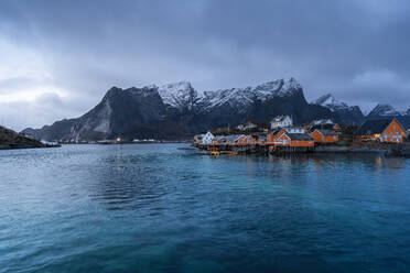 Häuser des Dorfes Reine am Meeresufer vor gefrorenen Bergen und bedecktem Himmel im Winter auf den Lofoten, Norwegen - ADSF33534