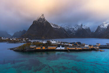 Das Dorf Reine liegt auf einer Insel, umgeben von ruhigem, blauem Wasser, schneebedeckten Bergen und bewölktem Himmel bei Sonnenuntergang - ADSF33539