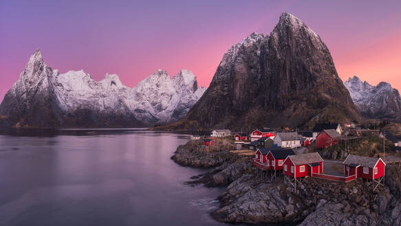 Atemberaubender Blick auf das Dorf Hamnoy mit seinen roten und weißen Häusern an der Küste des ruhigen Meeres vor dem Hintergrund des schneebedeckten Bergrückens und des Himmels bei Sonnenuntergang auf den Lofoten, Norwegen - ADSF33540