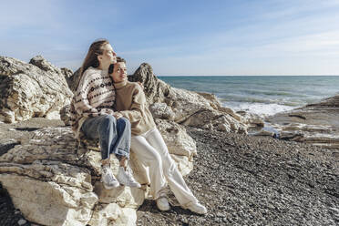 Mädchen sitzt auf einem Felsen und umarmt ihre Mutter am Strand - OMIF00523