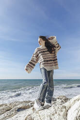 Teenager-Mädchen im Pullover steht auf einem Felsen am Strand - OMIF00524