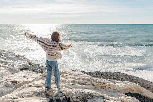 Teenager-Mädchen mit ausgestreckten Armen und Blick auf das Meer auf einem Felsen am Strand stehend - OMIF00526