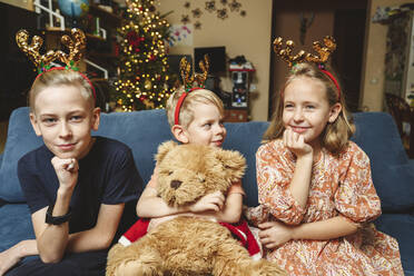 Siblings wearing reindeer antler sitting on sofa at Christmas - EYAF01903