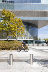 Man riding bicycle by modern office building on sunny day - IFRF01522