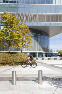 Mann auf dem Fahrrad neben einem modernen Bürogebäude an einem sonnigen Tag - IFRF01522